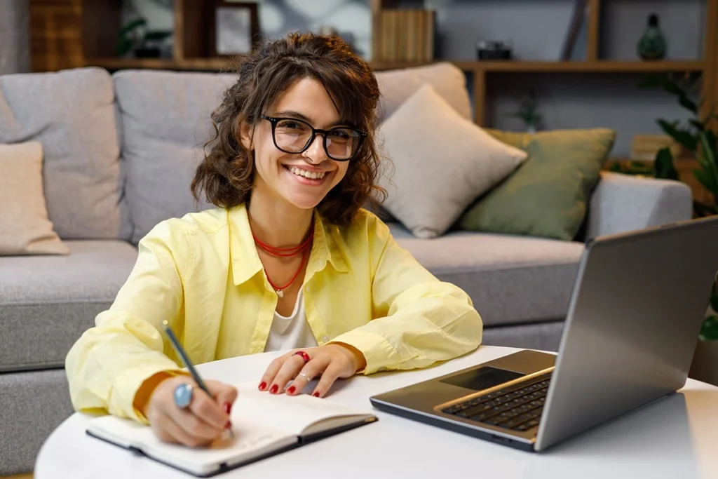 Curly-haired woman sitting on the floor in front of her couch with a laptop and notebook, smiling at the camera while engaging in self-paced training